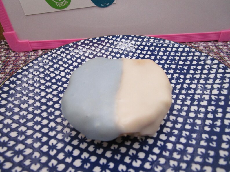 May include: A two-toned dessert, half light blue and half off-white, sits on a blue and white patterned plate. The dessert has a smooth, rounded shape. The plate's design features a repeating floral pattern.