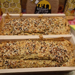 May include: Two loaves of bread in wooden baking molds, generously topped with a mix of seeds and spices. The bread is light brown, and the seeds are black and white. The loaves sit on a decorative plate.