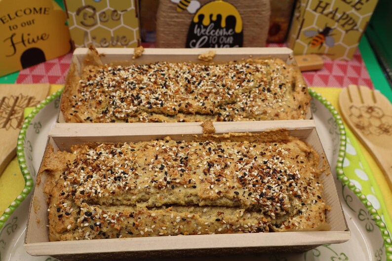 May include: Two loaves of bread in wooden baking molds, generously topped with a mix of seeds and spices. The bread is light brown, and the seeds are black and white. The loaves sit on a decorative plate.