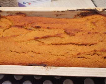 May include: A rectangular loaf of baked orange-colored bread or cake with a cracked, textured surface, contained within a light-colored wooden baking mold. The baked good features deep golden-brown edges and a porous, dense crumb structure.