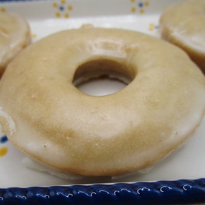May include: Close-up of glazed donuts. The donuts have a light brown color and a shiny glaze. The donuts are on a white plate with blue and yellow floral designs.
