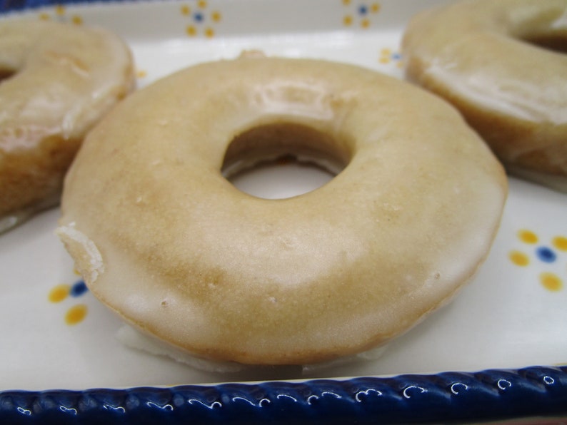 May include: Close-up of glazed donuts. The donuts have a light brown color and a shiny glaze. The donuts are on a white plate with blue and yellow floral designs.