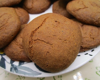 Könnte beinhalten: Eine Ansammlung von runden, braunen, gebackenen Keksen mit sichtbaren Rissen auf der Oberfläche, die auf einem weißen Teller mit einem dunklen Blattmuster angeordnet sind. Die Kekse haben ein dichtes, strukturiertes Aussehen und eine einheitliche braune Farbe.