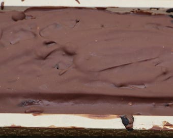 May include: A rectangular loaf of dark chocolate cake or bread encased in a light-colored wooden baking mold. The top surface is completely covered in a thick, smooth layer of milk chocolate ganache that extends to the edges of the container.
