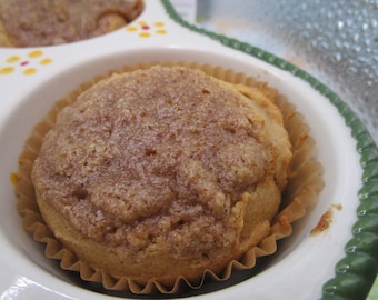 Può includere: Un muffin dorato con una copertura croccante e testurizzata di cannella e zucchero, inserito in un pirottino di carta beige. Il muffin è alloggiato in uno stampo in ceramica bianca con bordo smerlato verde e piccoli decori floreali gialli e rossi.