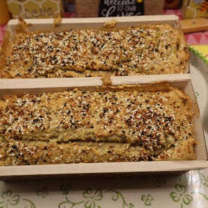 May include: Two loaves of bread in wooden baking pans, topped with a mix of seeds. The bread is light brown with a textured surface. The pans sit on a decorative platter with a floral design, alongside wooden utensils.