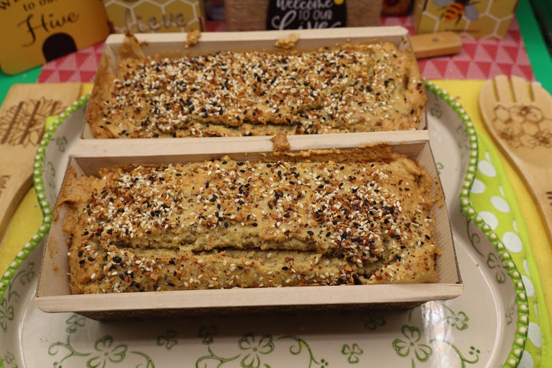 May include: Two loaves of bread in wooden baking pans, topped with a mix of seeds. The bread is light brown with a textured surface. The pans sit on a decorative platter with a floral design, alongside wooden utensils.