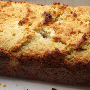 May include: A rectangular loaf of golden-brown baked bread with a textured, craggy crust. The loaf is positioned on an oval white serving platter that features small, decorative blue and yellow floral accents along its curved edges.