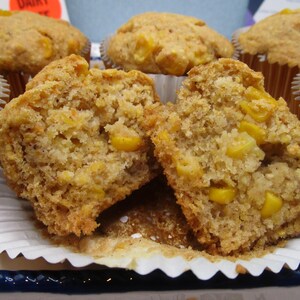 May include: Close-up of a corn muffin cut in half, revealing a moist interior with visible kernels. The muffin is golden brown with a slightly textured surface. Several other muffins are in the background, some with visible paper liners.