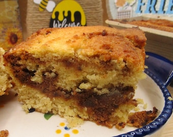 Könnte beinhalten: Ein dickes Stück goldbrauner Kuchen mit einem markanten, dunkelbraunen Zimt-Zucker-Wirbel in der Mitte. Der Kuchen hat eine krümelige, strukturierte Oberfläche und wird auf einem weißen Keramikt einem weißen Keramikteller mit blauem Wellenrand serviert.