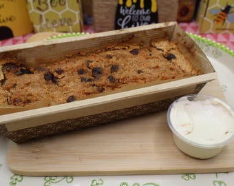 May include: A rectangular loaf of baked bread with visible dark inclusions, contained within a brown paper baking mould with a repeating pattern. A small, clear plastic container filled with white spread sits on a wooden board next to the loaf.
