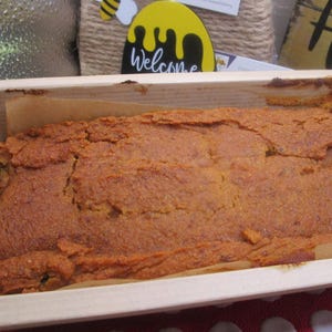 May include: A rectangular loaf of baked goods with a cracked, textured surface, presented in a wooden tray. The baked item is a warm, golden-brown color. A sign that says "Welcome" is visible in the background.