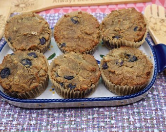 Könnte beinhalten: Sechs goldbraune Blaubeermuffins in Papierförmchen, angeordnet in einer rechteckigen weißen Keramik-Backform mit blauen Griffen. Die Form ist mit einem kleinen Blumenmuster verziert und von hellen Holzutensilien mit Gravuren umgeben.