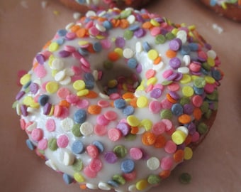 Könnte beinhalten: Ein runder, goldbrauner Donut mit einer glatten, weißen Glasur und einem großzügigen Belag aus kleinen, flachen, kreisförmigen Streuseln in verschiedenen Farben wie Pink, Orange, Gelb, Grün, Violett und Blau auf der gesamten Oberfläche.