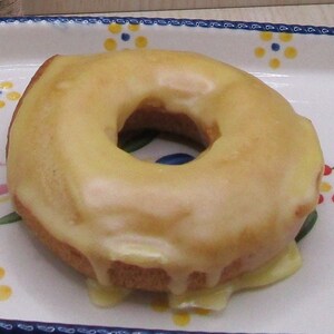 May include: A glazed donut with a yellow frosting sits on a decorative white plate. The plate has a blue rim and is decorated with pink, green, and yellow floral patterns. The donut's glaze is dripping down the sides.