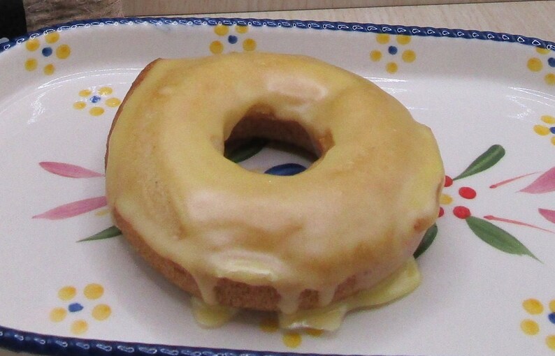 May include: A glazed donut with a yellow frosting sits on a decorative white plate. The plate has a blue rim and is decorated with pink, green, and yellow floral patterns. The donut's glaze is dripping down the sides.