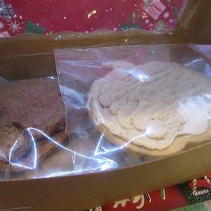 May include: Two cookies in clear plastic bags inside a brown cardboard box. One cookie is brown and the other is light brown.