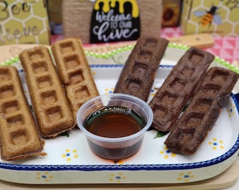 May include: Three golden-brown rectangular waffles and three dark brown chocolate rectangular waffles arranged on a white ceramic tray with blue and yellow floral accents, accompanied by a small clear plastic cup filled with amber syrup.