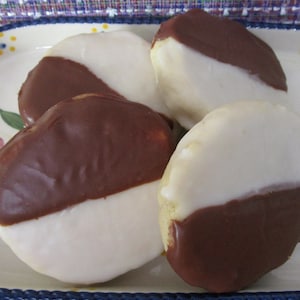 May include: Four black and white cookies on a decorative plate. Each cookie is oval-shaped, with one half covered in dark chocolate and the other half in white icing. The plate has a blue rim and floral designs.