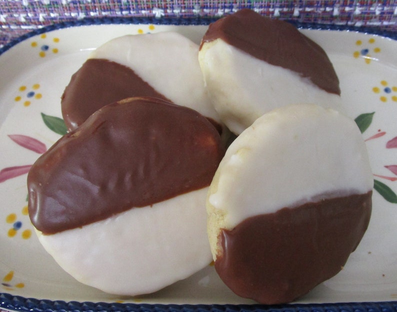 May include: Four black and white cookies on a decorative plate. Each cookie is oval-shaped, with one half covered in dark chocolate and the other half in white icing. The plate has a blue rim and floral designs.