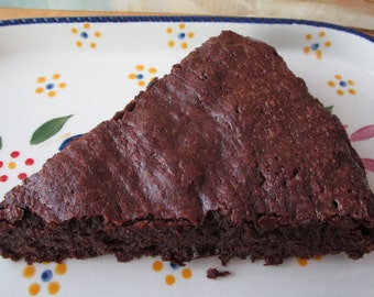 May include: A triangular slice of dark brown chocolate cake served on an oval ceramic platter. The platter features a white background with a hand-painted pattern of small yellow and blue flowers and green leaves along the rim.