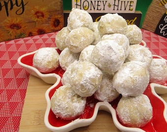 May include: A pile of snowballs cookies dusted with powdered sugar, presented on a red, star-shaped dish. The cookies are light in color and have a soft, rounded shape. The background includes a wooden sign with sunflowers.