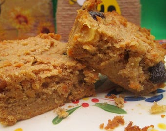 Könnte beinhalten: Zwei Scheiben Karottenkuchen mit Rosinen, serviert auf einem dekorativen Teller. Der Kuchen hat eine hellbraune Farbe mit sichtbaren Karottenstücken und Rosinen. Ein kleiner Behälter mit weißem Zuckerguss ist sichtbar.