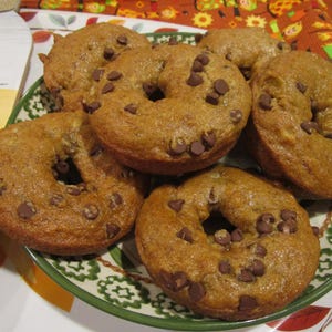 May include: A stack of golden-brown baked ring-shaped cakes topped with dark chocolate chips. The cakes are arranged on a round plate with a green rim and a decorative pattern, featuring a textured surface and a soft, porous crumb appearance.