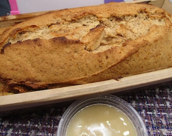 May include: A golden-brown loaf of baked bread sits inside a rectangular wooden baking mold. The crust has a cracked, textured surface with darker toasted edges. A small, clear plastic container filled with a light-colored liquid sauce sits nearby.