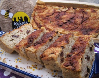 May include: Sliced loaf of bread with raisins and a cinnamon-sugar topping, presented on a decorative plate. A larger loaf is in the background, inside a wooden tray. A sign with a bee and the words "Welcome" is visible.