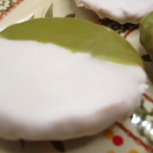 May include: Close-up of frosted cookies on a decorative plate. The cookies are round with a white base and a green glaze on top. The plate has a patterned rim with green and red accents.