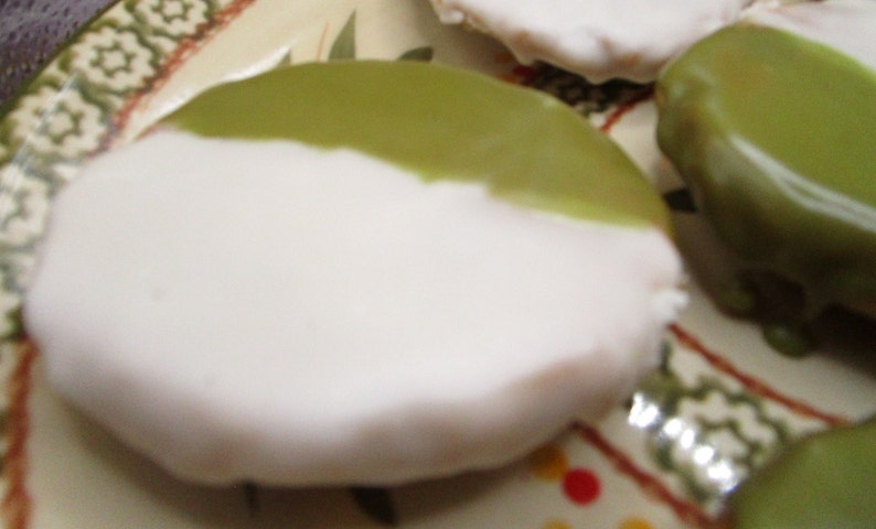 May include: Close-up of frosted cookies on a decorative plate. The cookies are round with a white base and a green glaze on top. The plate has a patterned rim with green and red accents.