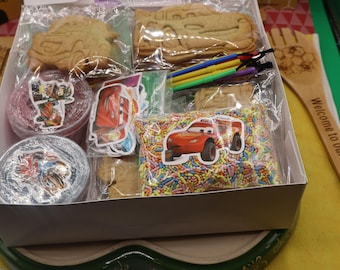 Puede incluir: Una caja de cartón blanco que contiene varios artículos, incluyendo galletas con forma de coche en bolsas de plástico, una bolsa de virutas de colores con una pegatina de coche de carreras rojo, recipientes pequeños, pegatinas y palitos.