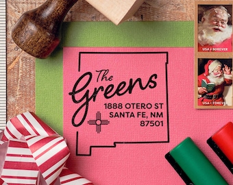 May include: A pink card with a black and white custom return address stamp. The stamp features the text "The Greens" in a cursive font, the address "1888 Otero St Santa Fe, NM 87501", and a stylized symbol of the state of New Mexico.