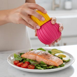 May include: A pink and yellow pepper grinder is held over a plate of food. The plate contains a cooked salmon fillet, fresh greens, sliced avocado, and cherry tomatoes. The pepper grinder is dispensing black pepper.