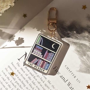 May include: A rectangular keychain with a bookshelf design under a night sky and a crescent moon. The keychain has a gold-coloured clasp and the words "THE READER" are printed on the front. The background includes a book with the title "Fire and Water".