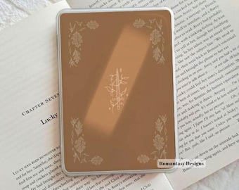 May include: A rectangular, light brown e-reader case featuring a thin, white line-art illustration of a sword entwined with vines in the centre, surrounded by a delicate border of oak leaves in each corner, with a clear, protective outer shell.
