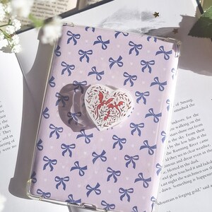 May include: A clear phone case with a lilac cover patterned with blue bows and white hearts. A heart-shaped pop socket with a red dragon design is attached. The image is on top of an open book.