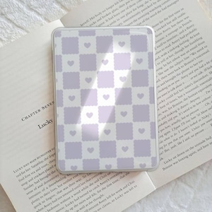 May include: A rectangular phone case featuring a repeating pattern of lavender squares and small white hearts. The case is displayed on an open book with text.