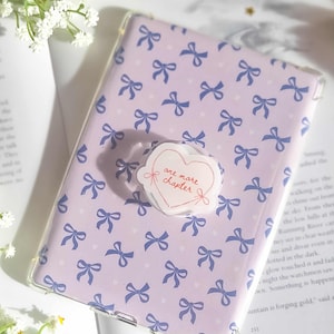 May include: A lilac phone case adorned with a pattern of blue bows and a heart-shaped phone grip. The grip features the text "one more chapter". The case is placed on an open book.