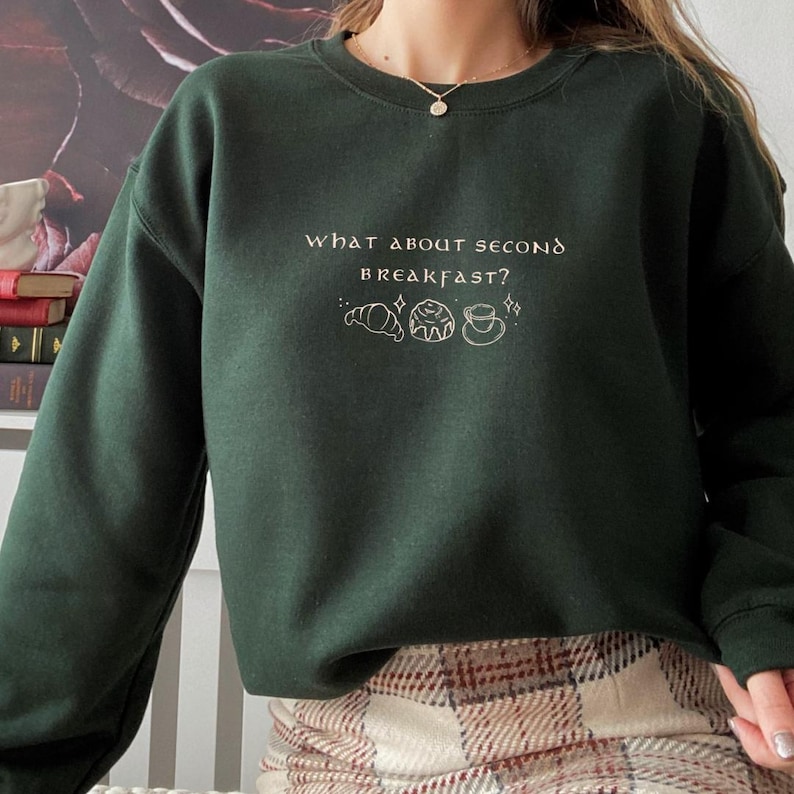 May include: A dark green sweatshirt with the text "what about second breakfast?" in white lettering. The sweatshirt features a small illustration of a croissant, a pastry, and a cup of coffee.
