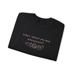 May include: Black t-shirt with the text "What about second breakfast?" in a light pink color. The text is accompanied by a small illustration of a croissant, a pastry, and a cup of coffee.