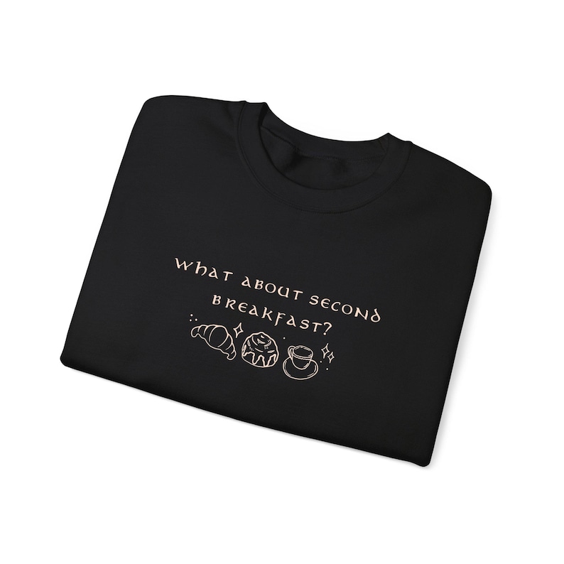May include: Black t-shirt with the text "What about second breakfast?" in a light pink color. The text is accompanied by a small illustration of a croissant, a pastry, and a cup of coffee.