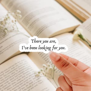 May include: A white sticker with the text "There you are, I've been looking for you" in black script. The sticker is held over an open book with a floral arrangement.