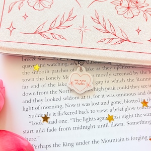 May include: A white book cover with red floral line art and a heart-shaped charm attached to the spine. The charm is white with a red heart outline and the text "one more chapter" in red. Small gold star confetti rests on the open book page.