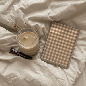May include: A clear glass tumbler filled with a beige liquid and a glass straw featuring a small brown bear charm, a dark brown tube labelled rhode, and a rectangular tablet case with a brown and white gingham pattern on the surface.