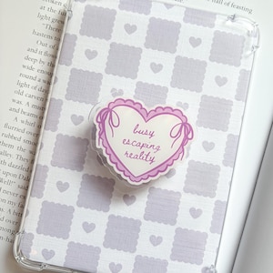 May include: Transparent phone case featuring a lavender and white checkered design with heart motifs. A heart-shaped pop socket with the words "busy escaping reality" is attached.