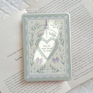 May include: A rectangular, pale green phone case with a decorative design. The design includes a sword, leaves, bows, and a heart with the text "born to read, forced to work."