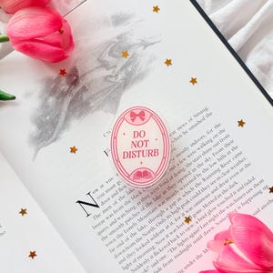 May include: An oval sticker with the text "DO NOT DISTURB" and a bow design, in pink and white, on an open book. Pink tulips and gold star-shaped confetti are scattered around the book.
