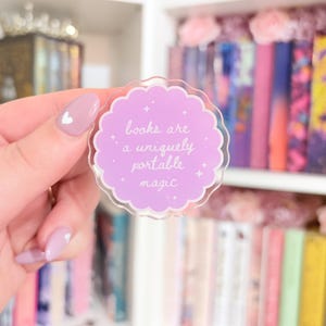 May include: A clear sticker with a scalloped edge and a light purple centre. The centre has the white text "books are a uniquely portable magic". Small white stars are scattered around the text. The sticker is held in front of a bookshelf.
